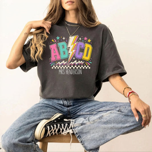 Person wearing a pepper t-shirt with colorful 'ABCD' design and jeans, sitting on a chair.