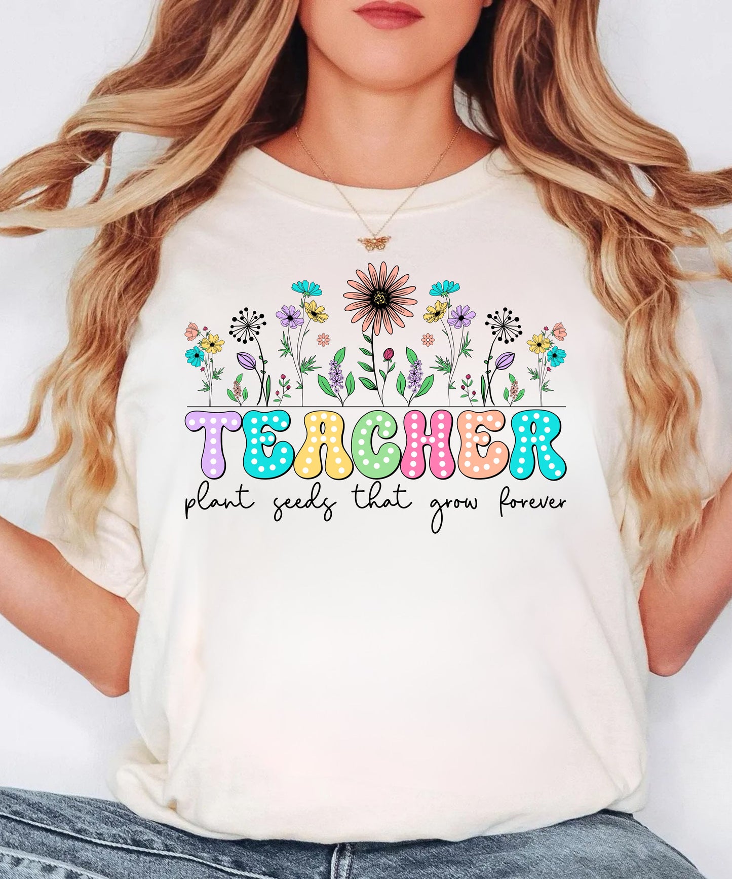 Person wearing an ivory t-shirt with colorful floral design and 'TEACHER plant seeds that grow forever' text.