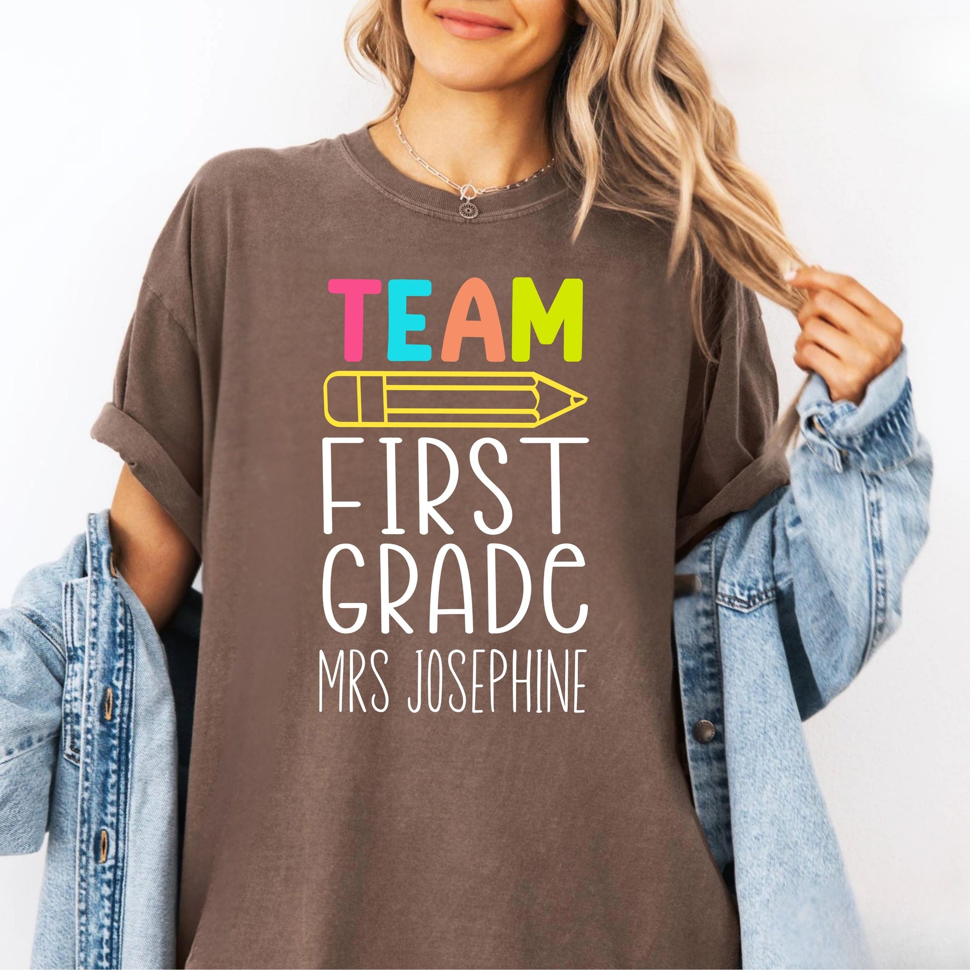 Person wearing an espresso colored  t-shirt with 'Team First Grade Mrs Josephine' text and colorful pencil graphic.