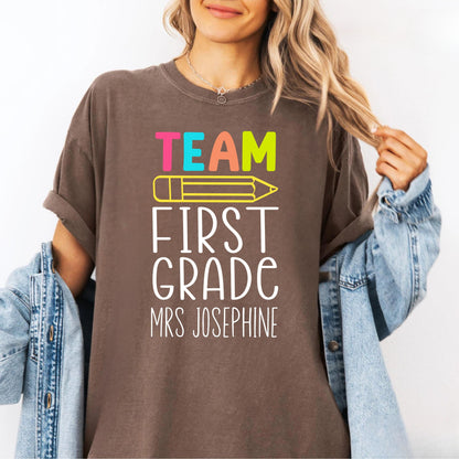 Person wearing an espresso colored  t-shirt with 'Team First Grade Mrs Josephine' text and colorful pencil graphic.