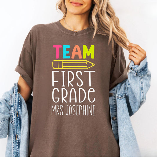Person wearing an espresso colored  t-shirt with 'Team First Grade Mrs Josephine' text and colorful pencil graphic.