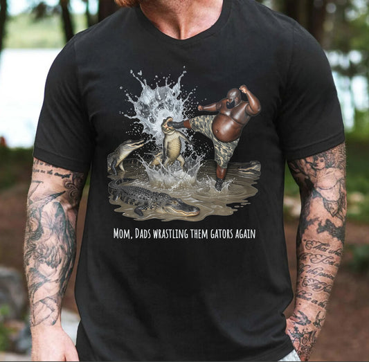 Black t-shirt with graphic design of one person wrestling alligators and text "mom, dad wrastling them gators again" worn by a person with tattoo