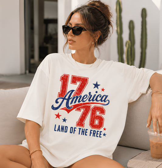 Woman wearing a white t-shirt with '17 America 76 Land of the Free' design, sitting on a couch.