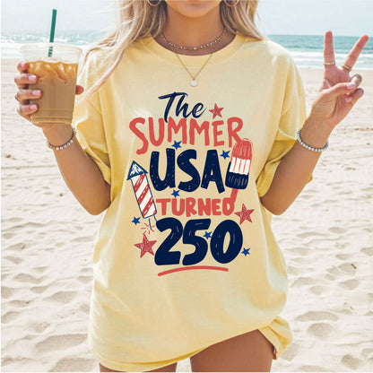 Person on a beach wearing a butter t-shirt with 'The Summer USA Turned 250' text.