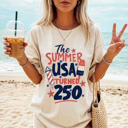 Woman on a beach wearing an ivory colored t shirt 'The Summer USA Turned 250', holding a drink.