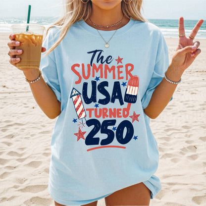 Person on a beach wearing a chambray blue t-shirt with 'The Summer USA Turned 250' text.