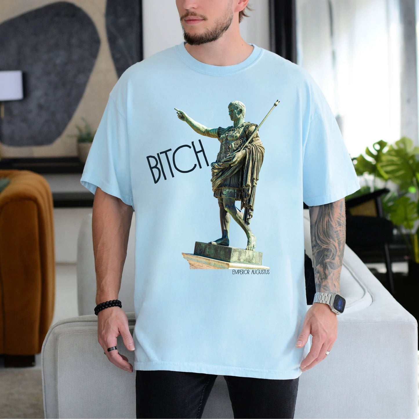 person wearing a chambray blue  t-shirt with a graphic of a statue pointing and the word 'Bitch' on a white background