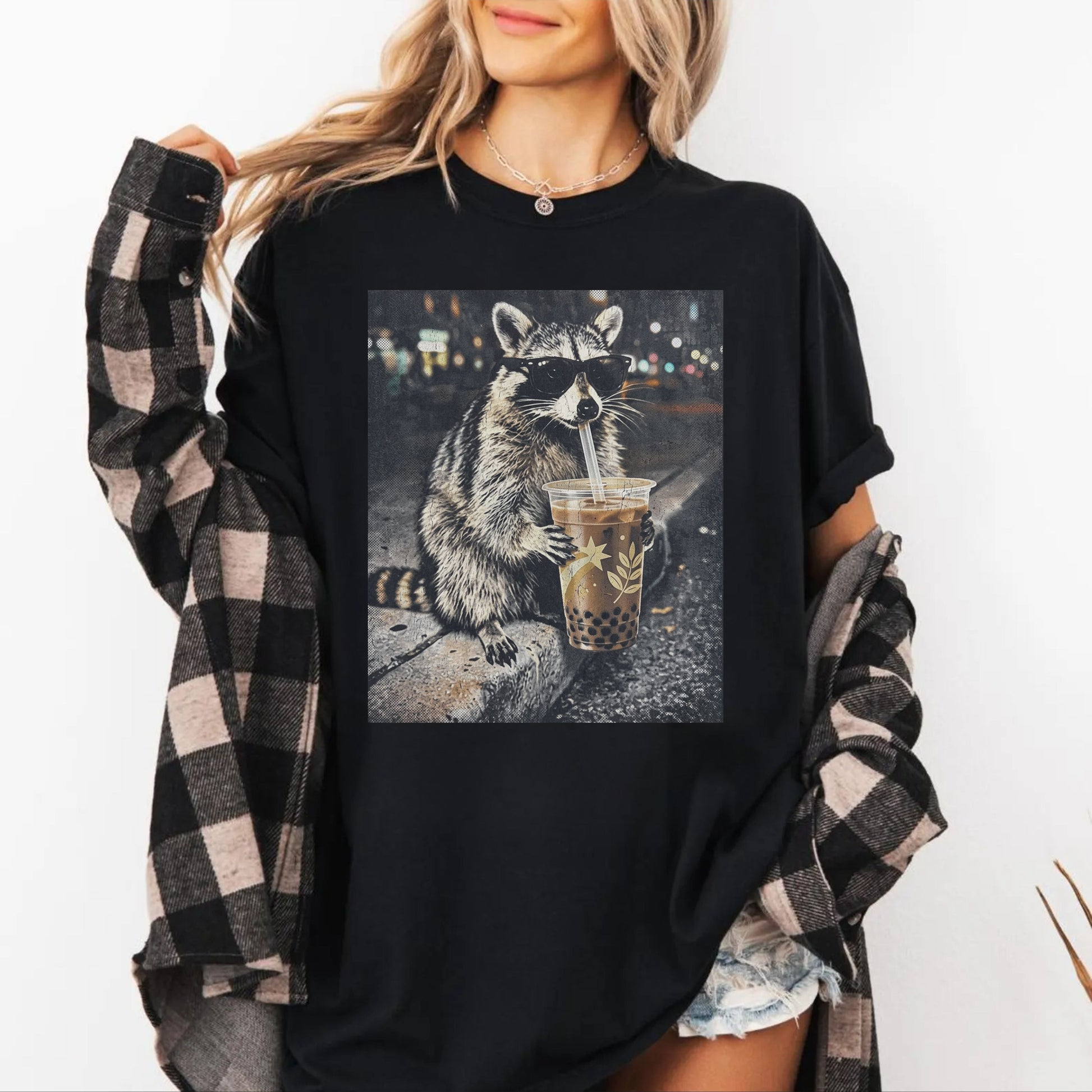Person wearing a black t-shirt with a raccoon graphic holding a cup of bobba tea design, plaid shirt over shoulders.