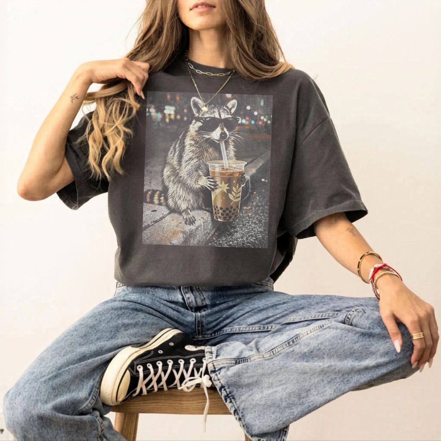 Person wearing a pepper t-shirt with a graphic of a raccoon holding a drink, sitting on a chair.