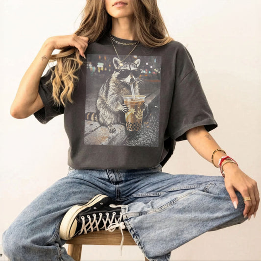 Person wearing a pepper t-shirt with a graphic of a raccoon holding a drink, sitting on a chair.