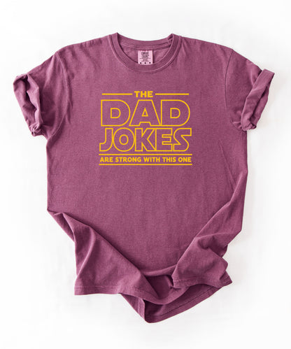a berry t-shirt with yellow text 'The Dad Jokes Are Strong With This One' in front of a blurred background.