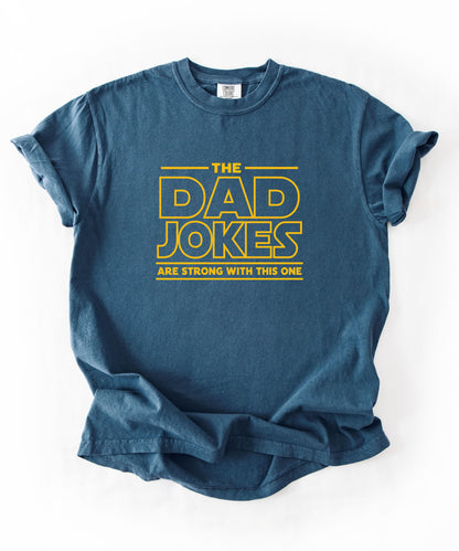 a midnight t-shirt with yellow text 'The Dad Jokes Are Strong With This One' in front of a blurred background.
