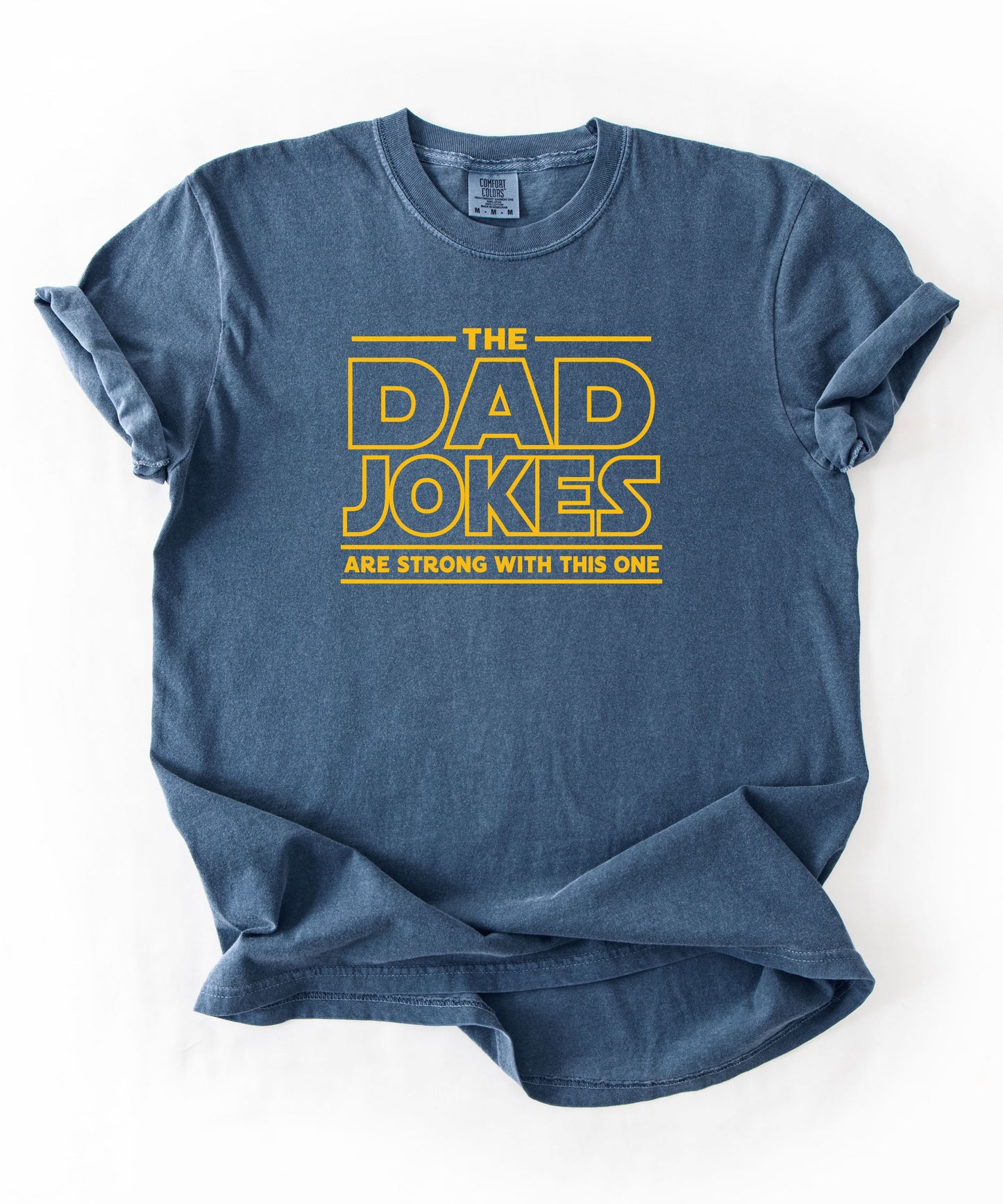 a denim t-shirt with yellow text 'The Dad Jokes Are Strong With This One' in front of a blurred background.