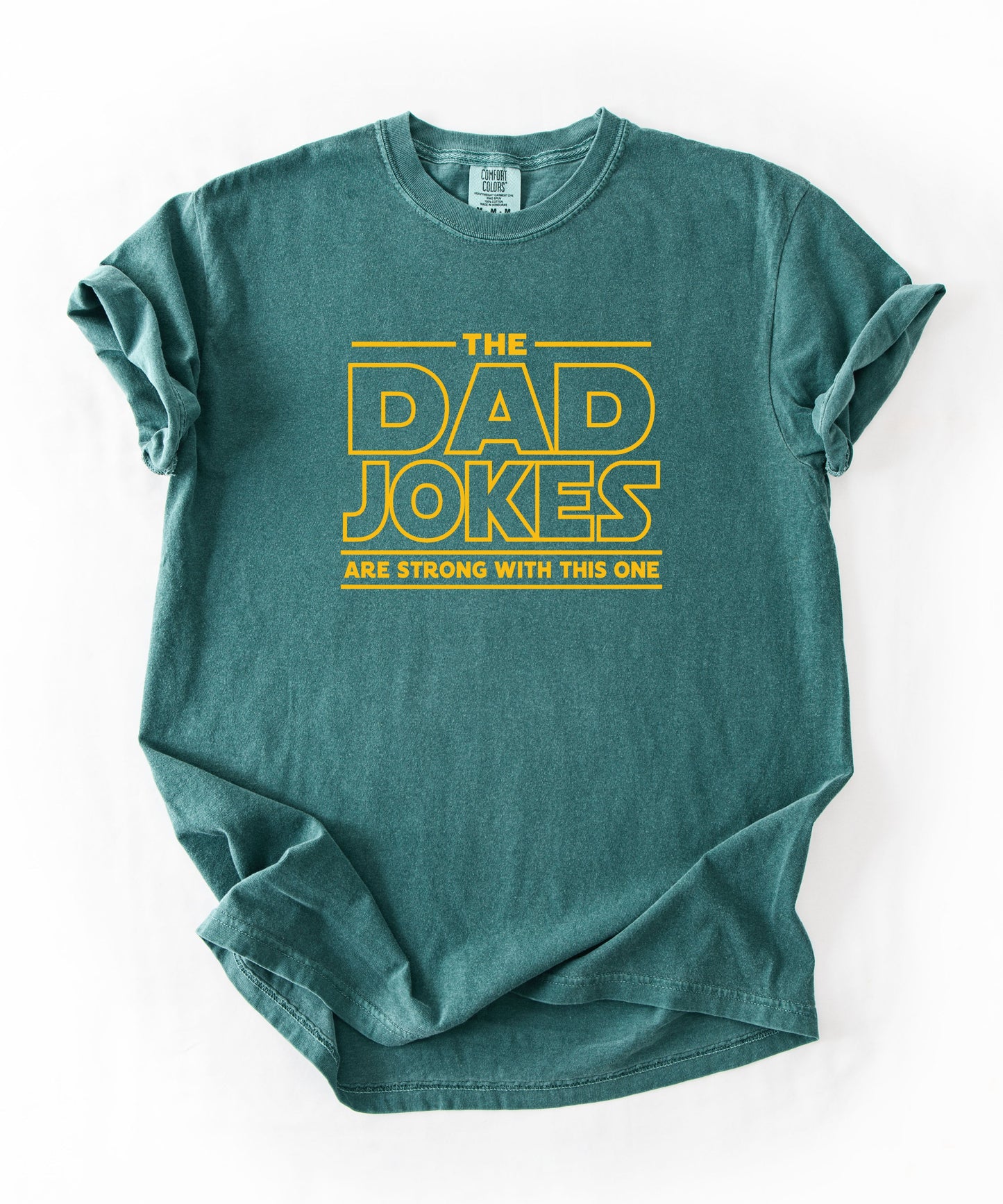 a blue spruce t-shirt with yellow text 'The Dad Jokes Are Strong With This One' in front of a blurred background.