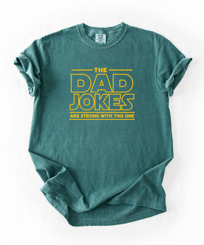 a blue spruce t-shirt with yellow text 'The Dad Jokes Are Strong With This One' in front of a blurred background.