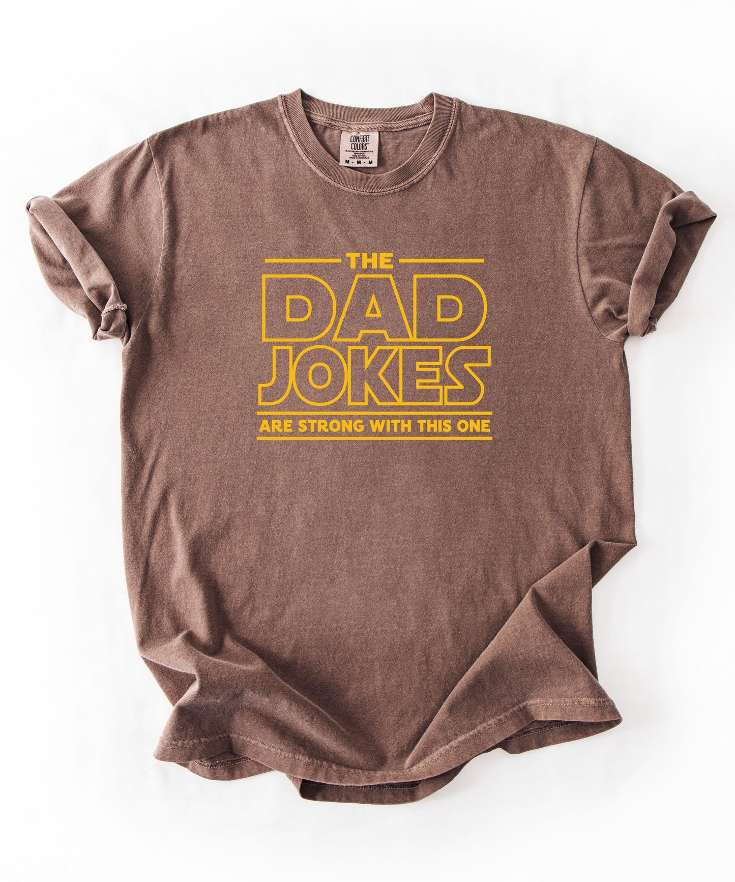 an espresso t-shirt with yellow text 'The Dad Jokes Are Strong With This One' in front of a blurred background.