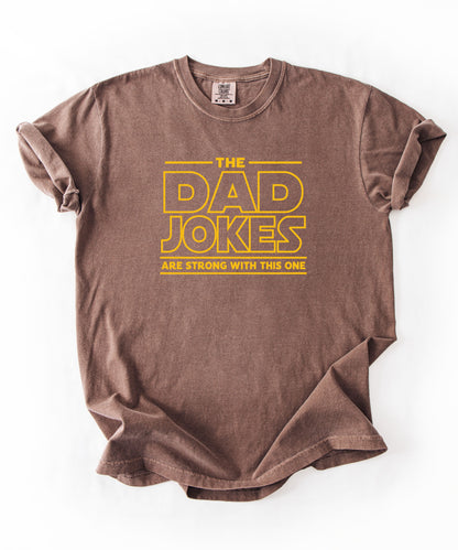an espresso t-shirt with yellow text 'The Dad Jokes Are Strong With This One' in front of a blurred background.