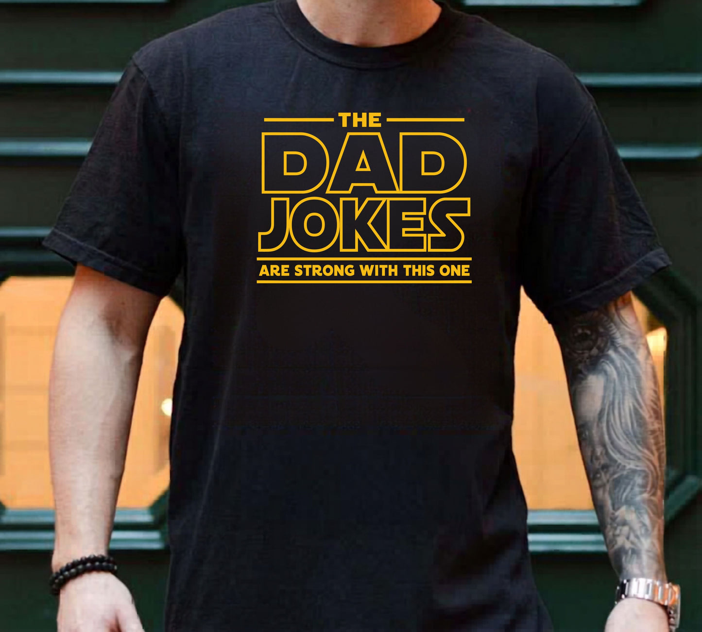 Person wearing a black t-shirt with yellow text 'The Dad Jokes Are Strong With This One' in front of a blurred background.