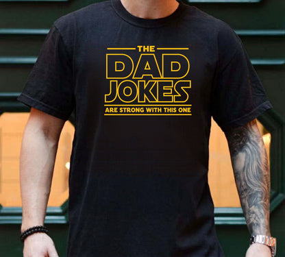 Person wearing a black t-shirt with yellow text 'The Dad Jokes Are Strong With This One' in front of a blurred background.