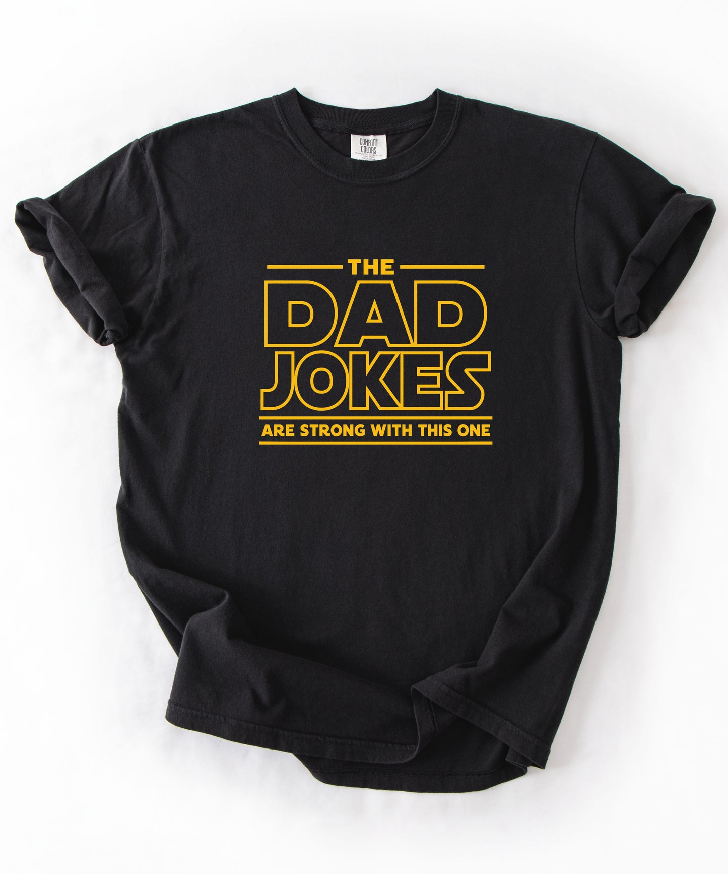 a black t-shirt with yellow text 'The Dad Jokes Are Strong With This One' in front of a blurred background.