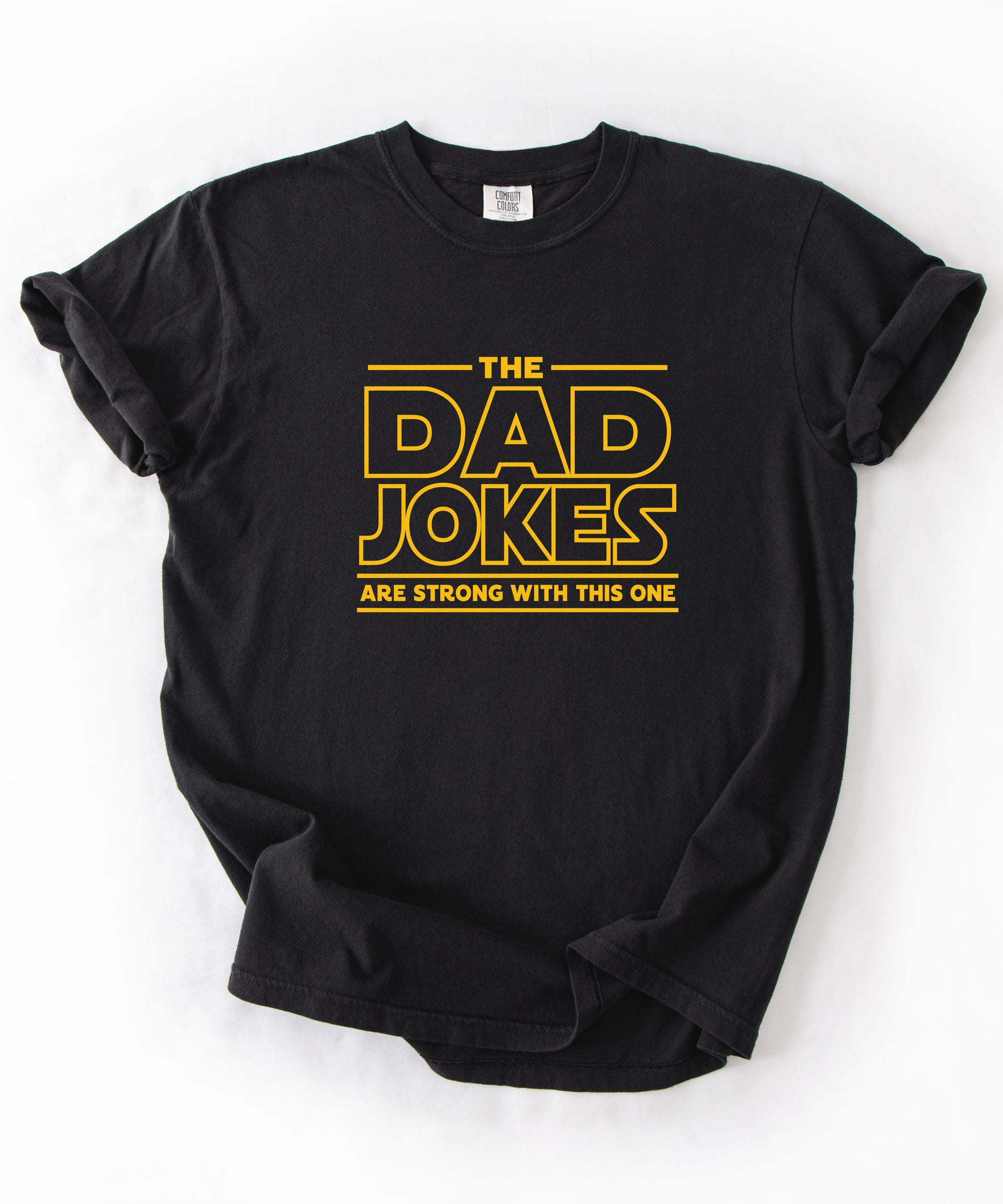 a black t-shirt with yellow text 'The Dad Jokes Are Strong With This One' in front of a blurred background.