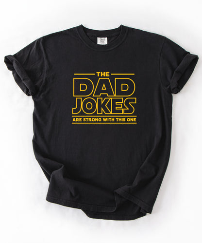 a black t-shirt with yellow text 'The Dad Jokes Are Strong With This One' in front of a blurred background.