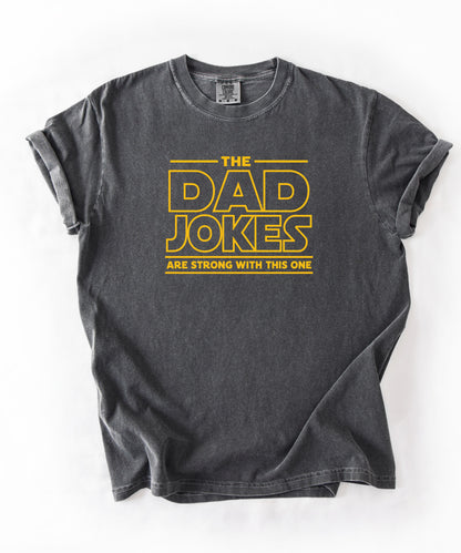 a pepper t-shirt with yellow text 'The Dad Jokes Are Strong With This One' in front of a blurred background.