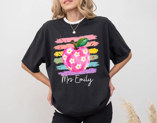Personalized teacher t-shirt in Black with floral apple graphic and colorful brushstroke stripes, custom name in script — garment-dyed cotton tee