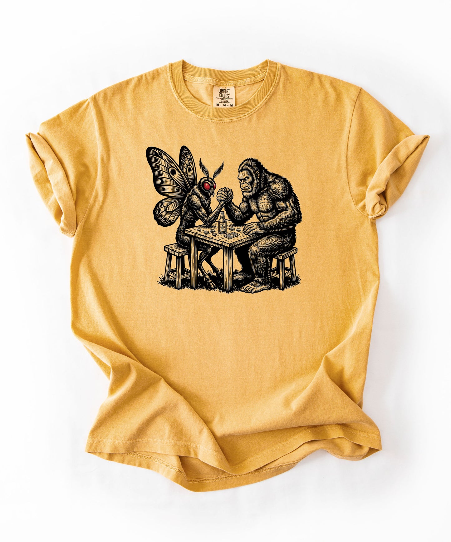 Mustard garment-dyed unisex Bigfoot vs Mothman arm wrestling graphic tee — funny cryptid shirt