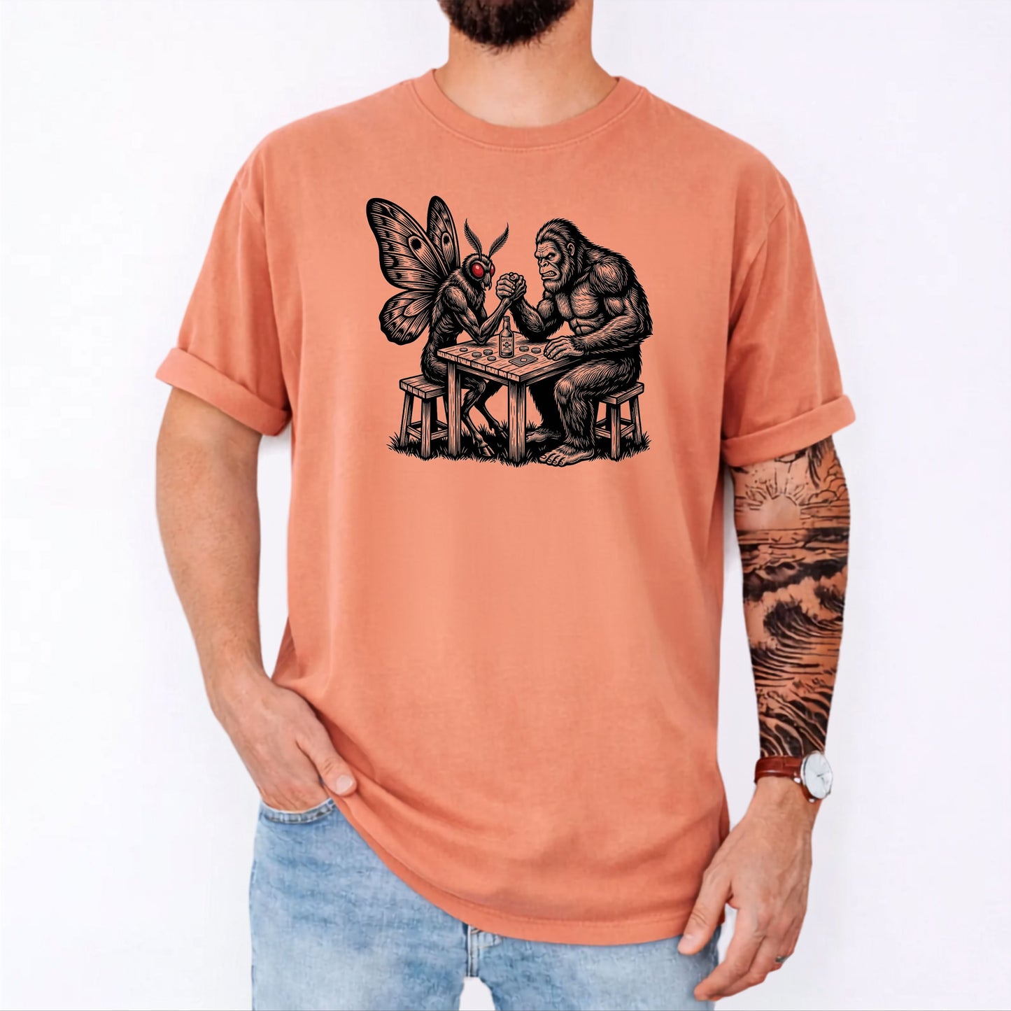 Terracotta garment-dyed unisex Bigfoot vs Mothman arm wrestling graphic tee — funny cryptid shirt