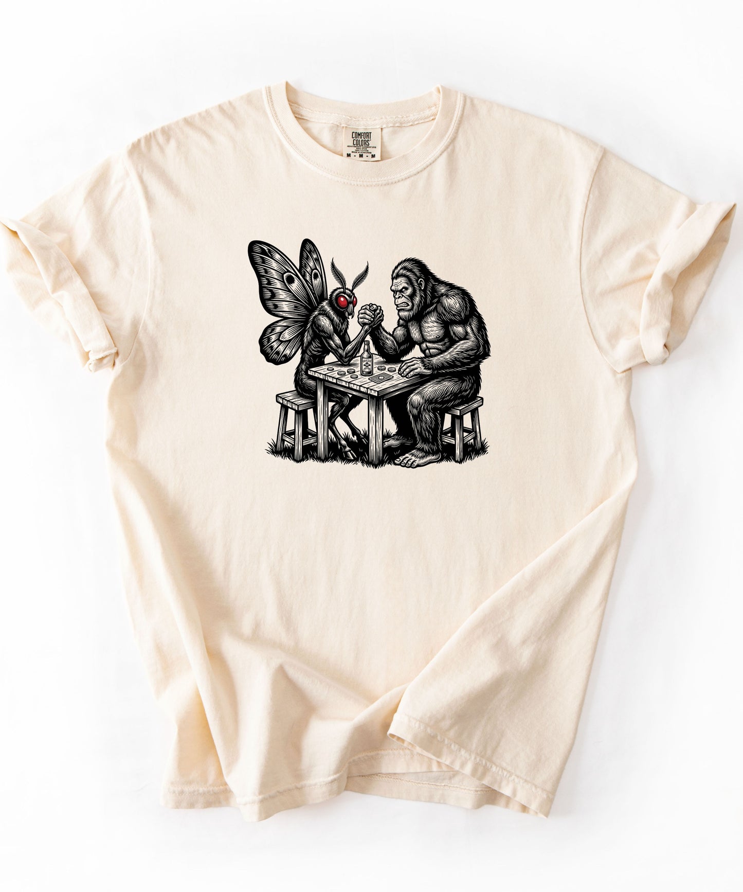 Ivory garment-dyed unisex Bigfoot vs Mothman arm wrestling graphic tee — funny cryptid shirt