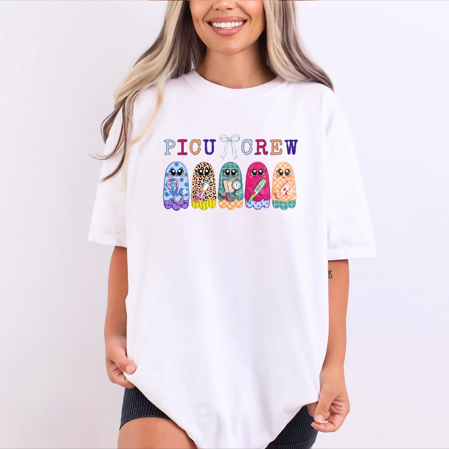 Woman wearing White garment-dyed PICU Crew t-shirt with five colorful patterned ghosts holding nursing tools — stethoscope, syringe, blood pressure cuff, thermometer, and bandaids