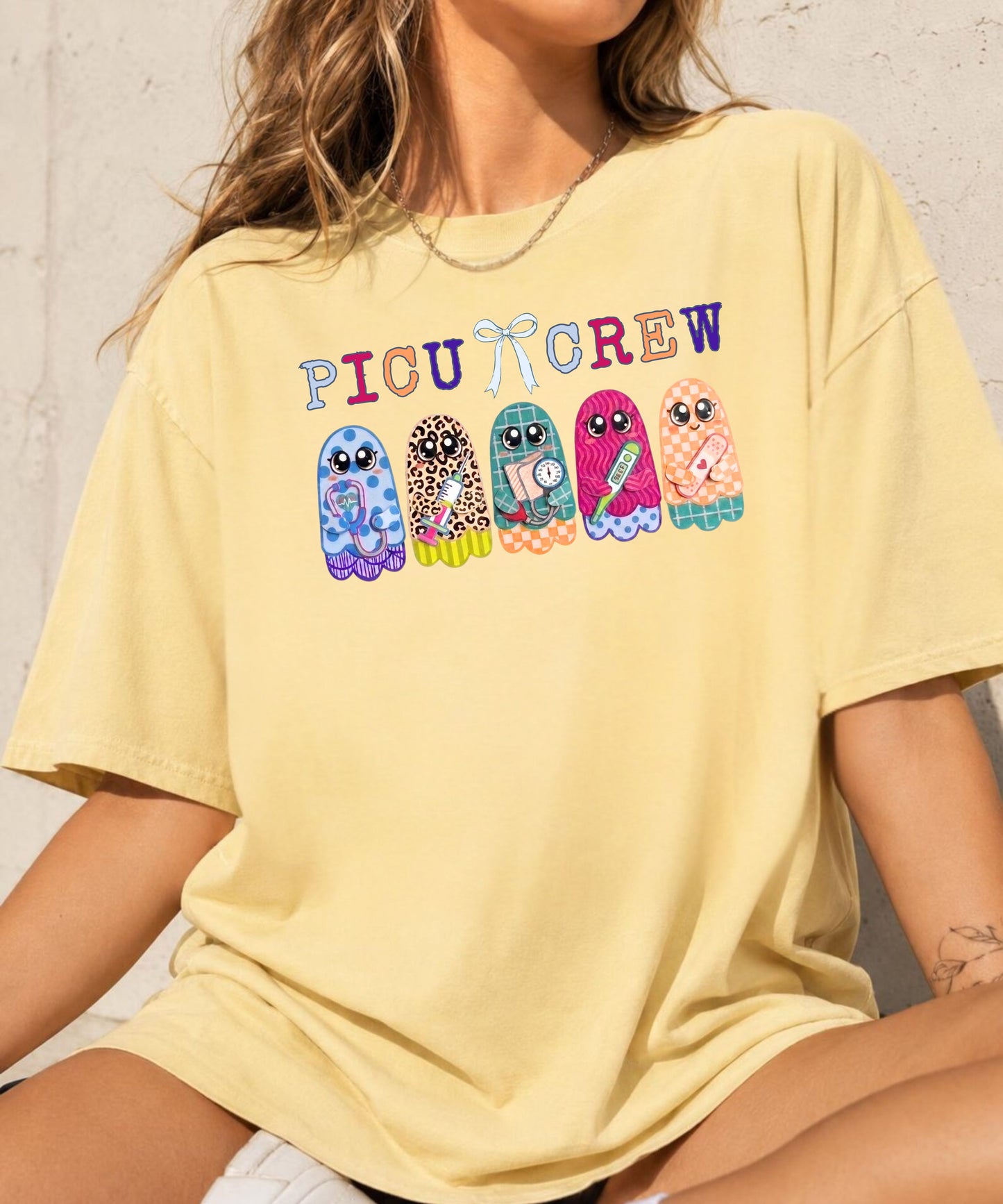 Woman wearing butter yellow garment-dyed PICU Crew t-shirt with five colorful patterned ghosts holding nursing tools — stethoscope, syringe, blood pressure cuff, thermometer, and bandaids