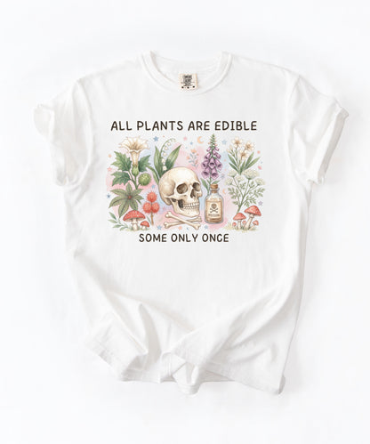 A white short-sleeve t-shirt with a vintage-style botanical graphic. The graphic features a central skull surrounded by illustrated mushrooms, flowers, and a small apothecary bottle, with the text 'ALL PLANTS ARE EDIBLE' above and 'SOME ONLY ONCE' below.