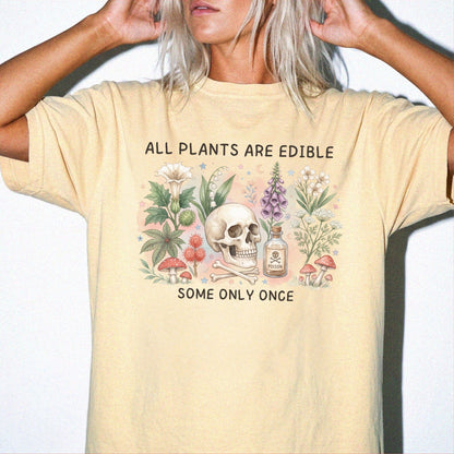 A woman wearing a butter yellow graphic t-shirt with the text 'ALL PLANTS ARE EDIBLE SOME ONLY ONCE' and a central illustration of a skull surrounded by plants and mushrooms.