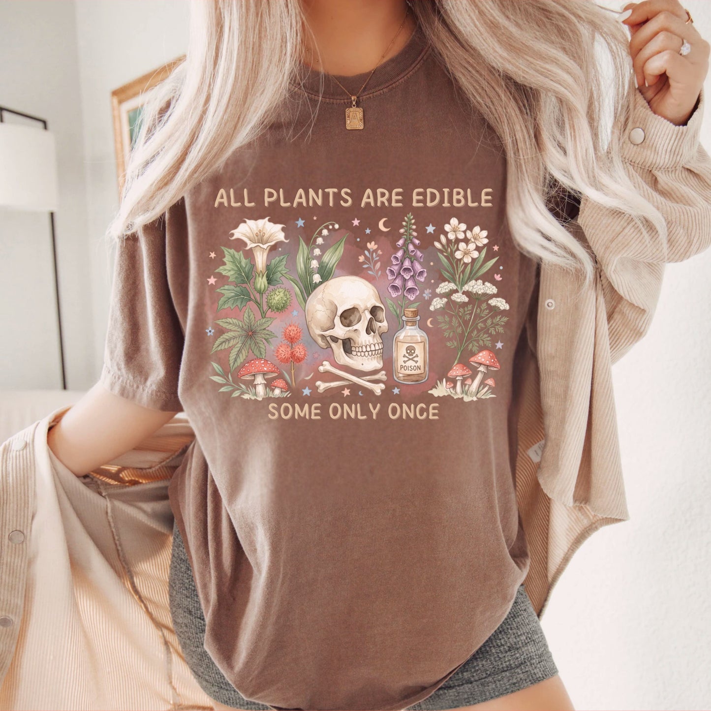 A woman wearing an espresso colored graphic t-shirt with a skull, flowers, and the text 'ALL PLANTS ARE EDIBLE SOME ONLY ONCE', styled with an open beige button-up shirt and gray shorts.