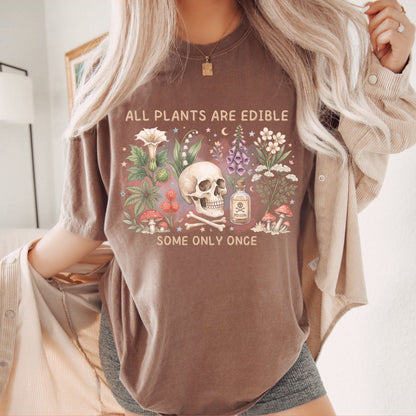 A woman wearing an espresso colored graphic t-shirt with a skull, flowers, and the text 'ALL PLANTS ARE EDIBLE SOME ONLY ONCE', styled with an open beige button-up shirt and gray shorts.