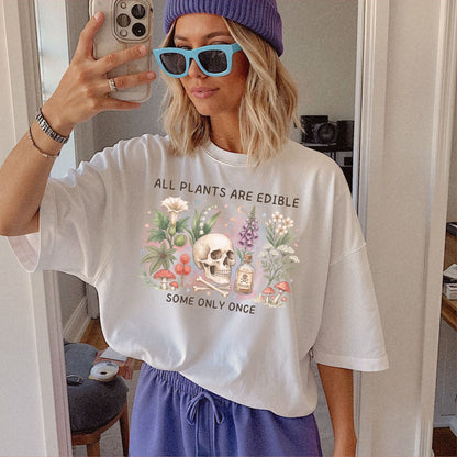 A woman takes a mirror selfie wearing an oversized white graphic T-shirt with a skull and flowers and the text 'ALL PLANTS ARE EDIBLE SOME ONLY ONCE', paired with a purple beanie, light blue sunglasses, and purple sweatpants.