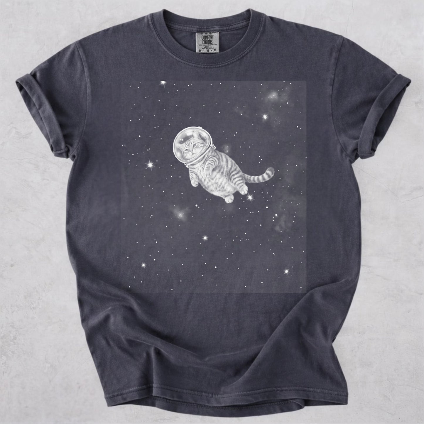 A Graphite gray crewneck t-shirt featuring a white line-art graphic of a cat floating in space with stars, laid flat on a light gray textured background.