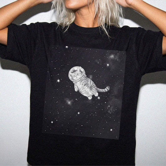 A person wearing a black short-sleeve T-shirt with a large square graphic on the chest showing a gray tabby cat floating in space with stars.
