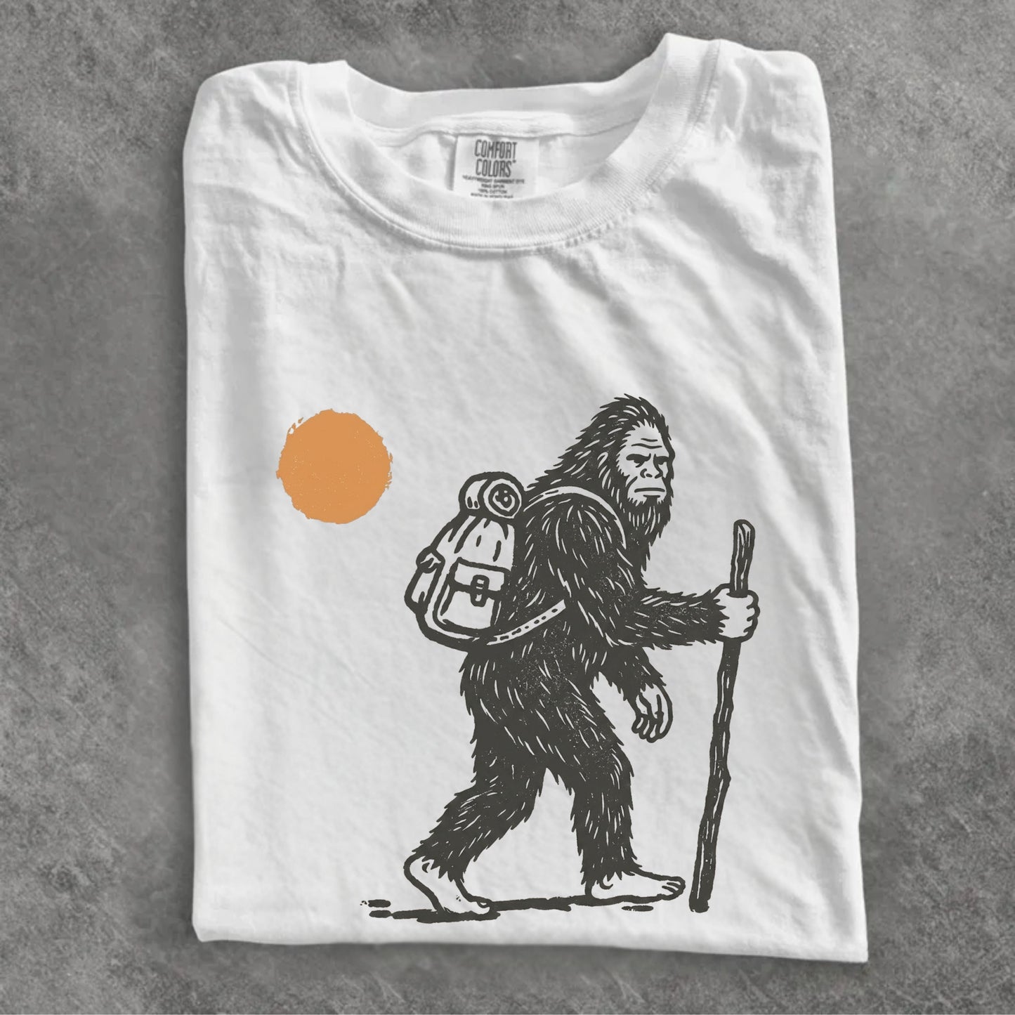 A folded white t-shirt with a black line art graphic of a Bigfoot hiker holding a staff, next to an orange sun, displayed on a textured gray background.