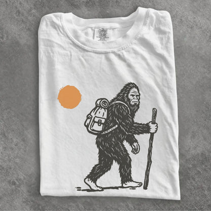 A folded white t-shirt with a black line art graphic of a Bigfoot hiker holding a staff, next to an orange sun, displayed on a textured gray background.
