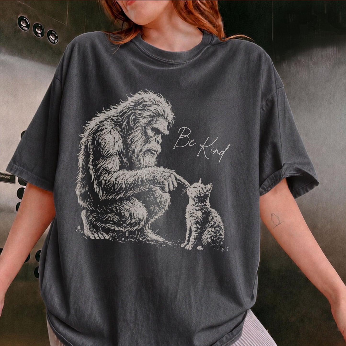 A person wearing an oversized Pepper graphic t-shirt. The t-shirt features a detailed black and white illustration of a Bigfoot figure reaching down to a kitten, with the handwritten text 'Be Kind' printed next to it.