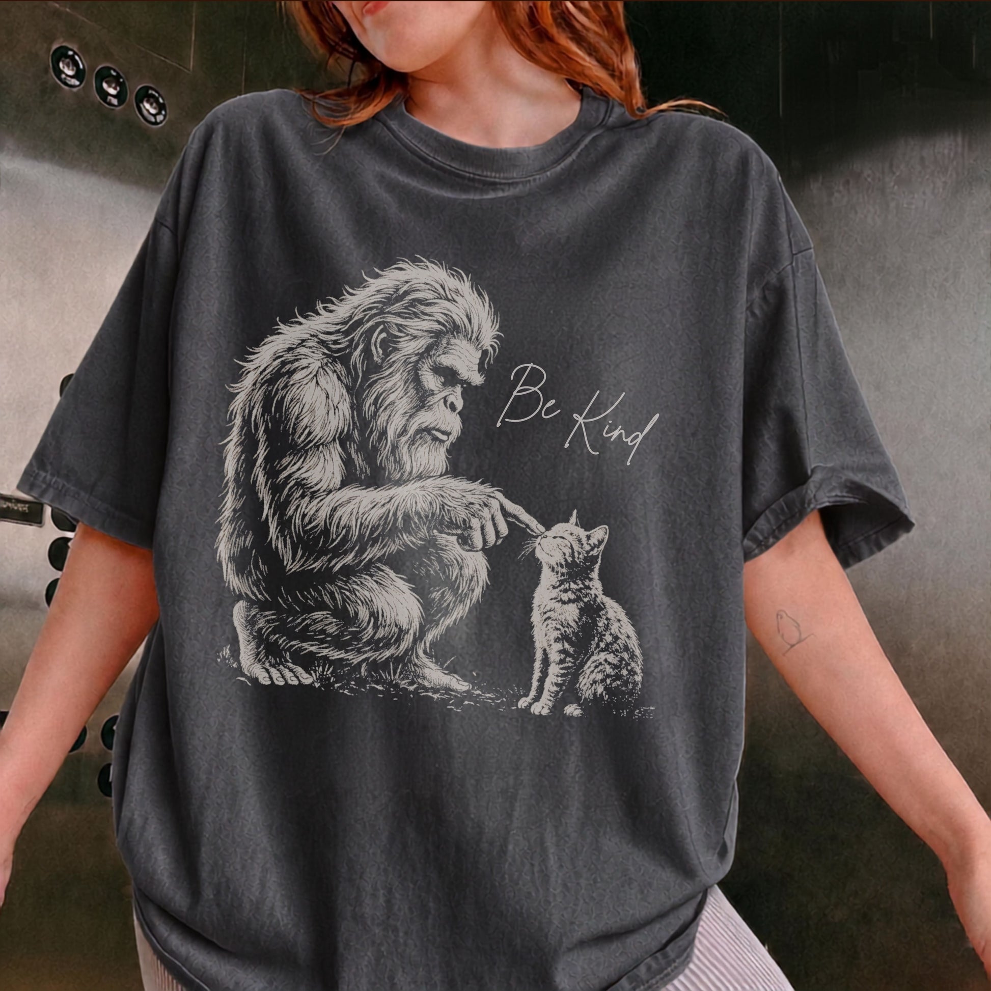 A person wearing an oversized Pepper graphic t-shirt. The t-shirt features a detailed black and white illustration of a Bigfoot figure reaching down to a kitten, with the handwritten text 'Be Kind' printed next to it.