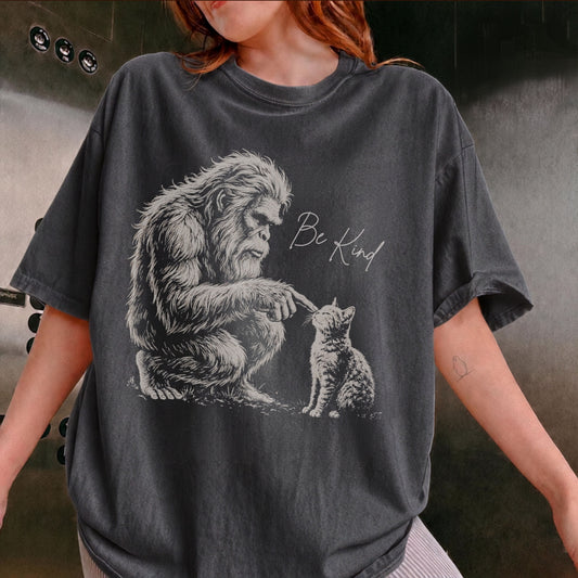 A person wearing an oversized Pepper graphic t-shirt. The t-shirt features a detailed black and white illustration of a Bigfoot figure reaching down to a kitten, with the handwritten text 'Be Kind' printed next to it.