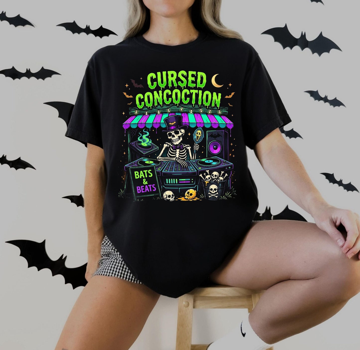 A woman wearing a black graphic t-shirt with a colorful 'Cursed Concoction' bat-themed print, sitting on a wooden stool against a light gray wall decorated with bat silhouettes.