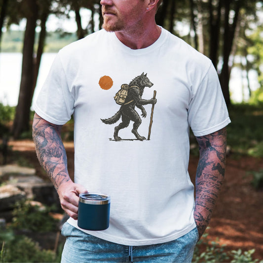 A man wearing a white graphic t-shirt with a hiking wolf design, standing outdoors in a wooded area and holding a dark blue insulated tumbler.