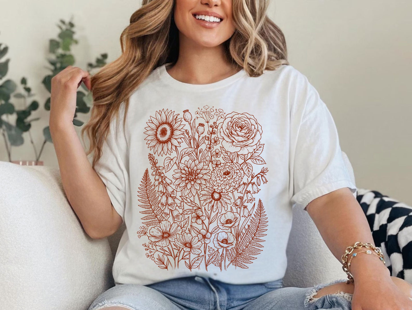 A woman wearing a white short-sleeve t-shirt with a rust-colored floral line art print on the front, paired with light-wash ripped jeans, sitting on a white couch.