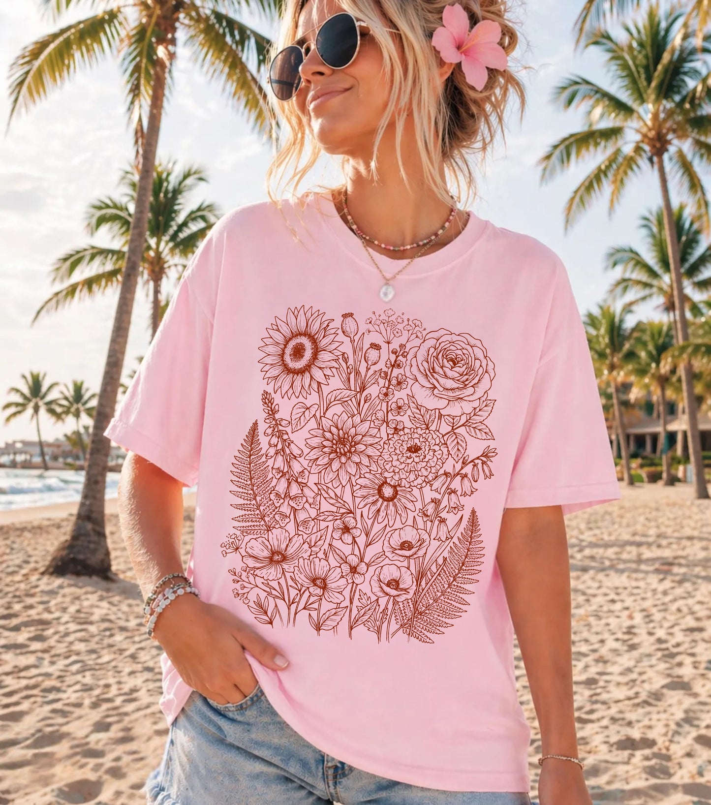 A woman wearing a Blossom pink oversized t-shirt with a brown floral line art print, standing on a sandy beach with palm trees in the background.