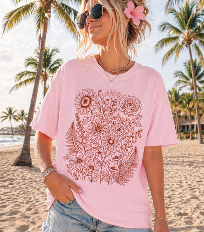 A woman wearing a Blossom pink oversized t-shirt with a brown floral line art print, standing on a sandy beach with palm trees in the background.